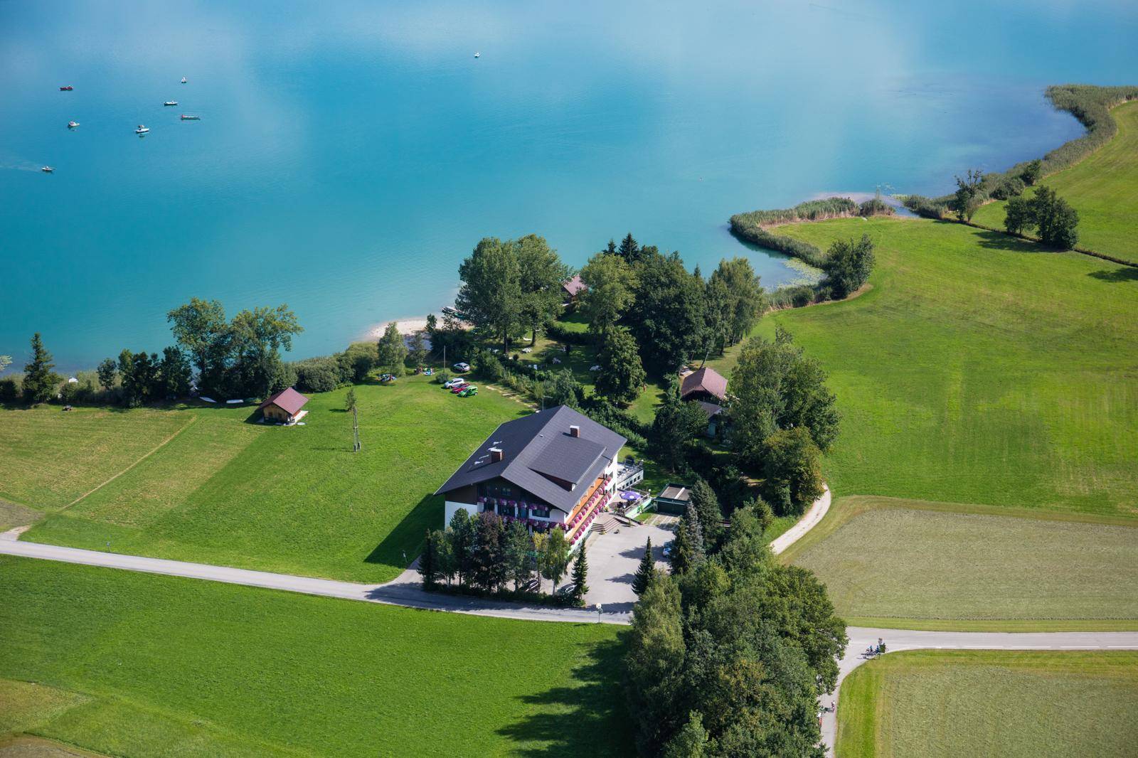 Nahaufnahme aus der Luft eines Hotels am See mit umliegenden Wiesen, Bäumen und einem Parkplatz.