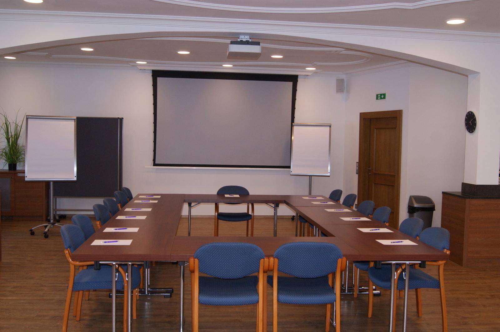 Konferenzraum mit U-förmiger Tischordnung, Projektionsleinwand und Präsentationsmaterialien für Meetings.