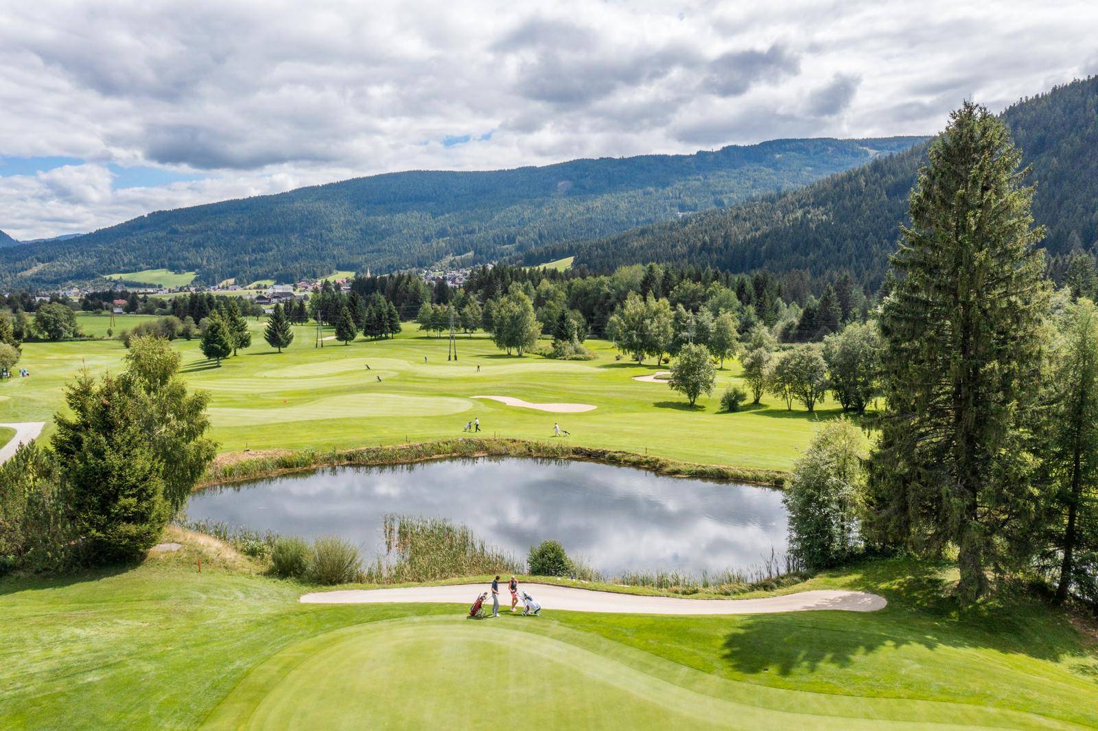 Luftaufnahme eines Golfplatzes mit einem Teich, umgeben von Bäumen und grünen Hügeln.
