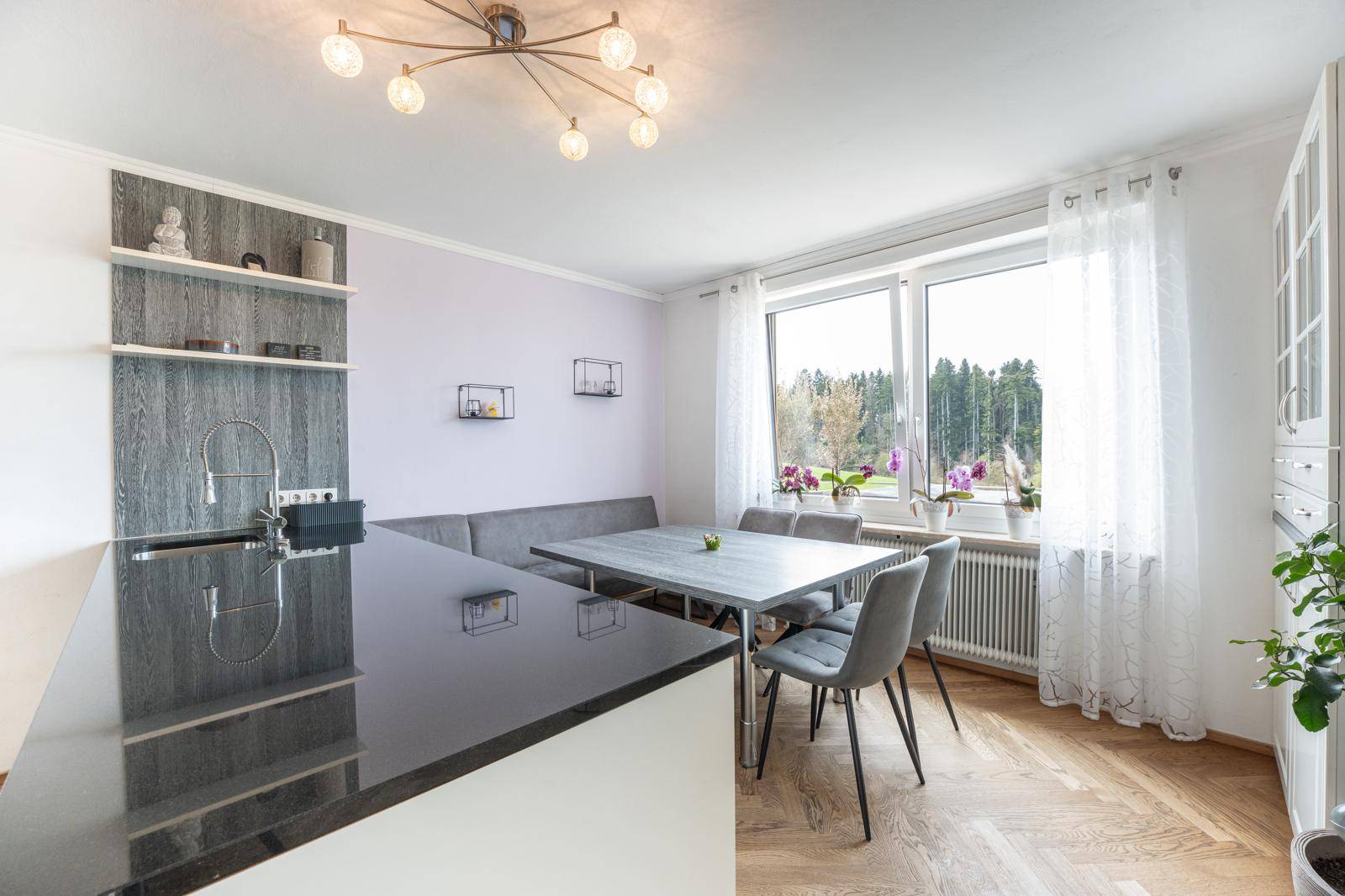 Moderne Küche mit dunkler Arbeitsplatte, Essbereich und großem Fenster mit Blick ins Grüne.