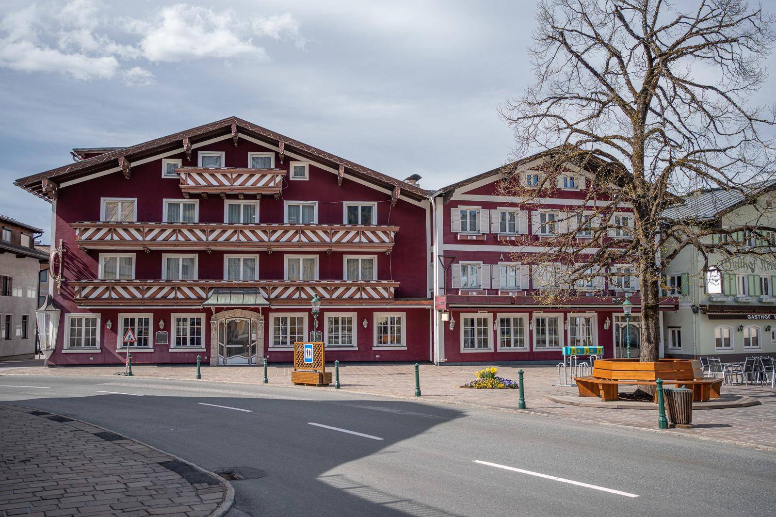 Gesamtansicht des roten Gebäudes und des Dorfplatzes mit einer Bank und einem kahlen Baum im Frühling.