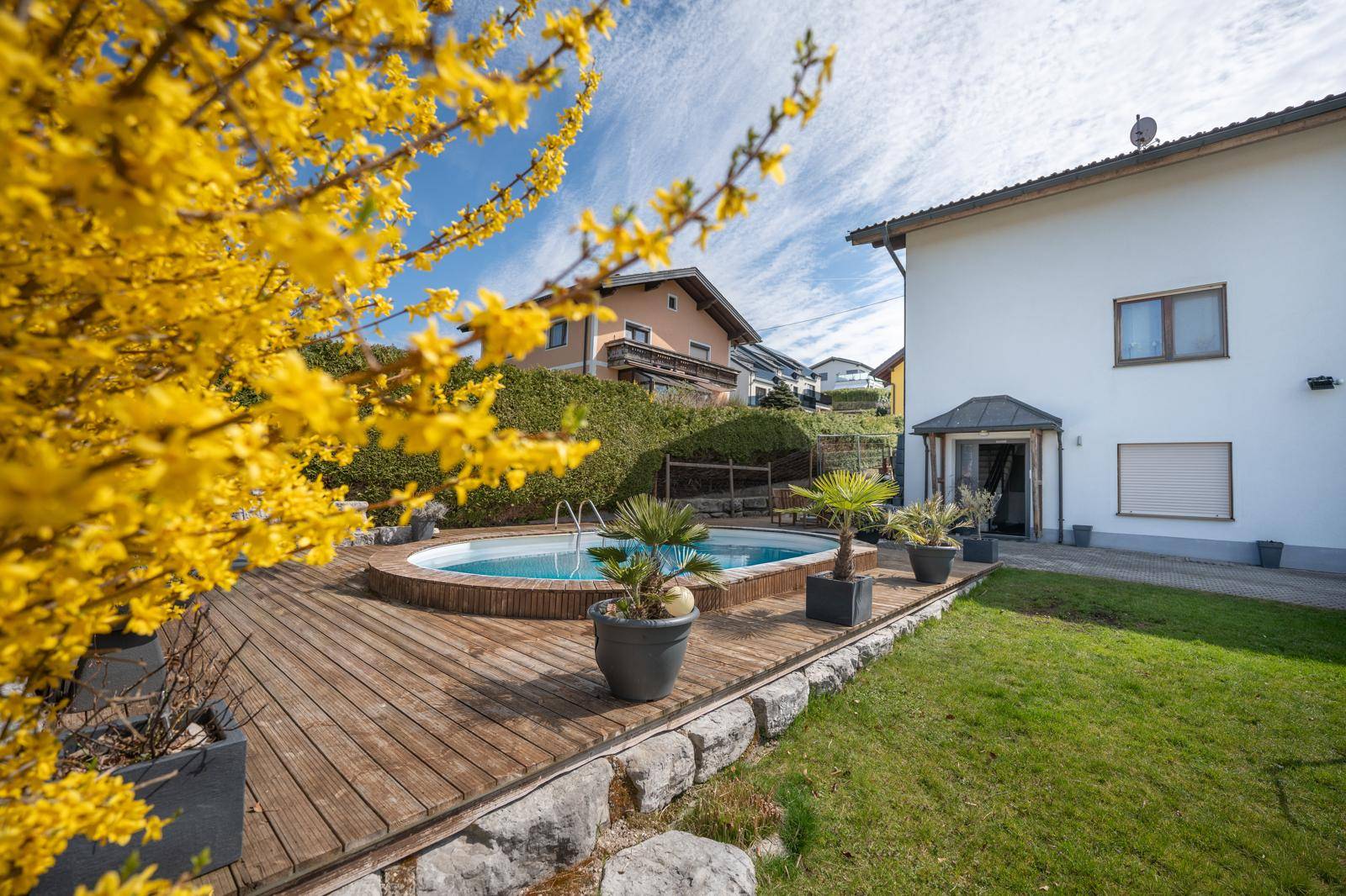 Runder Pool mit Holzdeck und Palmen im Garten, umgeben von blühenden Sträuchern unter blauem Himmel.