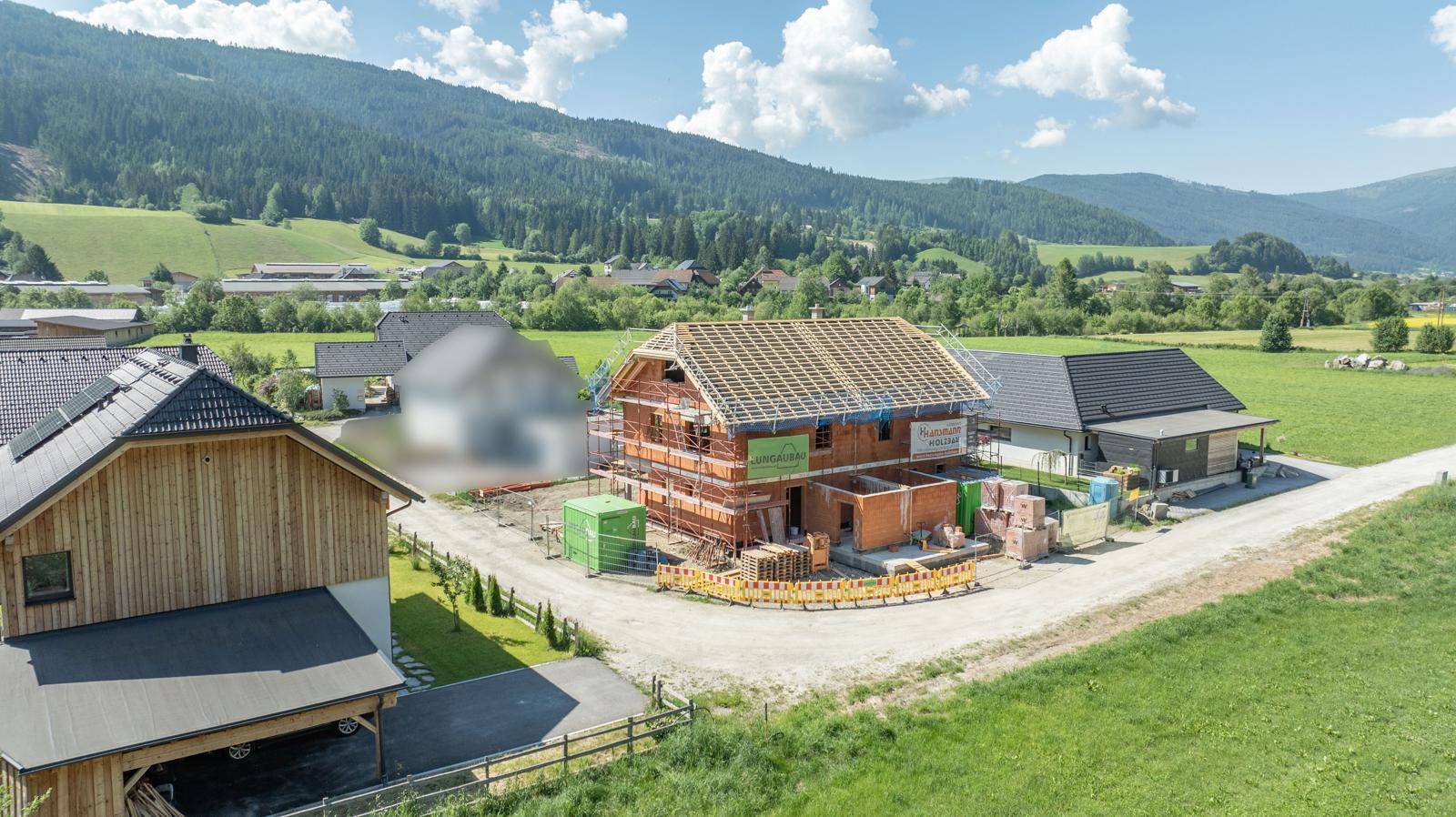 Baustelle eines Einfamilienhauses im Rohbau mit Dachstuhl, umgeben von grünen Feldern.