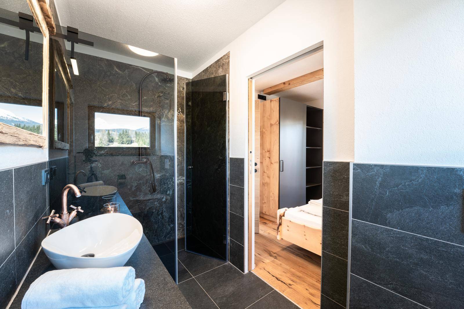 Stilvolles Badezimmer mit dunklen Fliesen, begehbarer Dusche und Blick auf das Schlafzimmer.