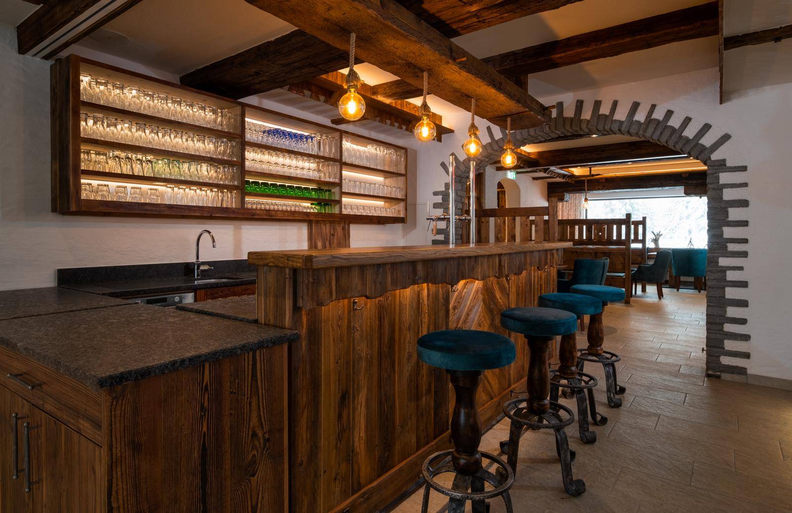 Rustikale Bar mit Holztresen, Hockern, beleuchteten Regalen und freiliegenden Deckenbalken.