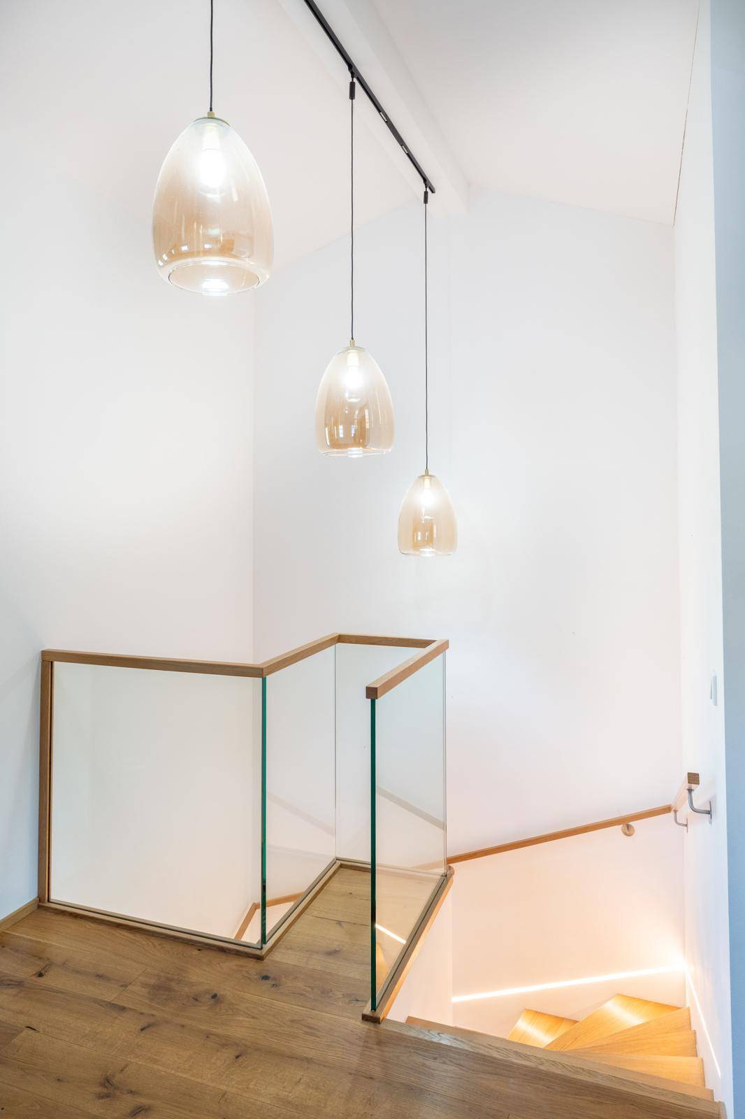 Modernes Treppenhaus mit Holzstufen, Glasgeländer und stilvollen Hängelampen.