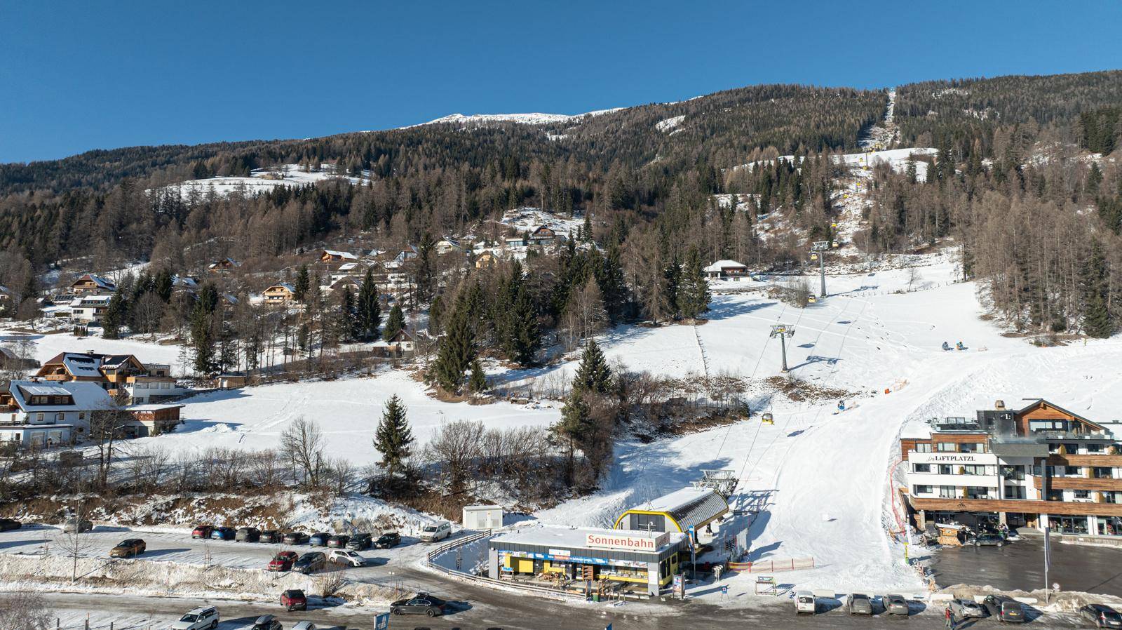 Winterlandschaft mit Skilift und Pisten, umgeben von verschneiten Bergen und einem Hotelgebäude.