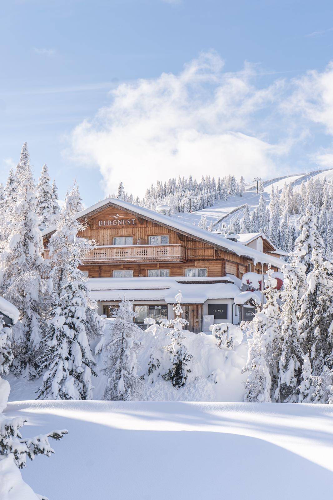 Traditionelles Holzgebäude im Winter, umgeben von schneebedeckten Bäumen und Bergen unter blauem Himmel.