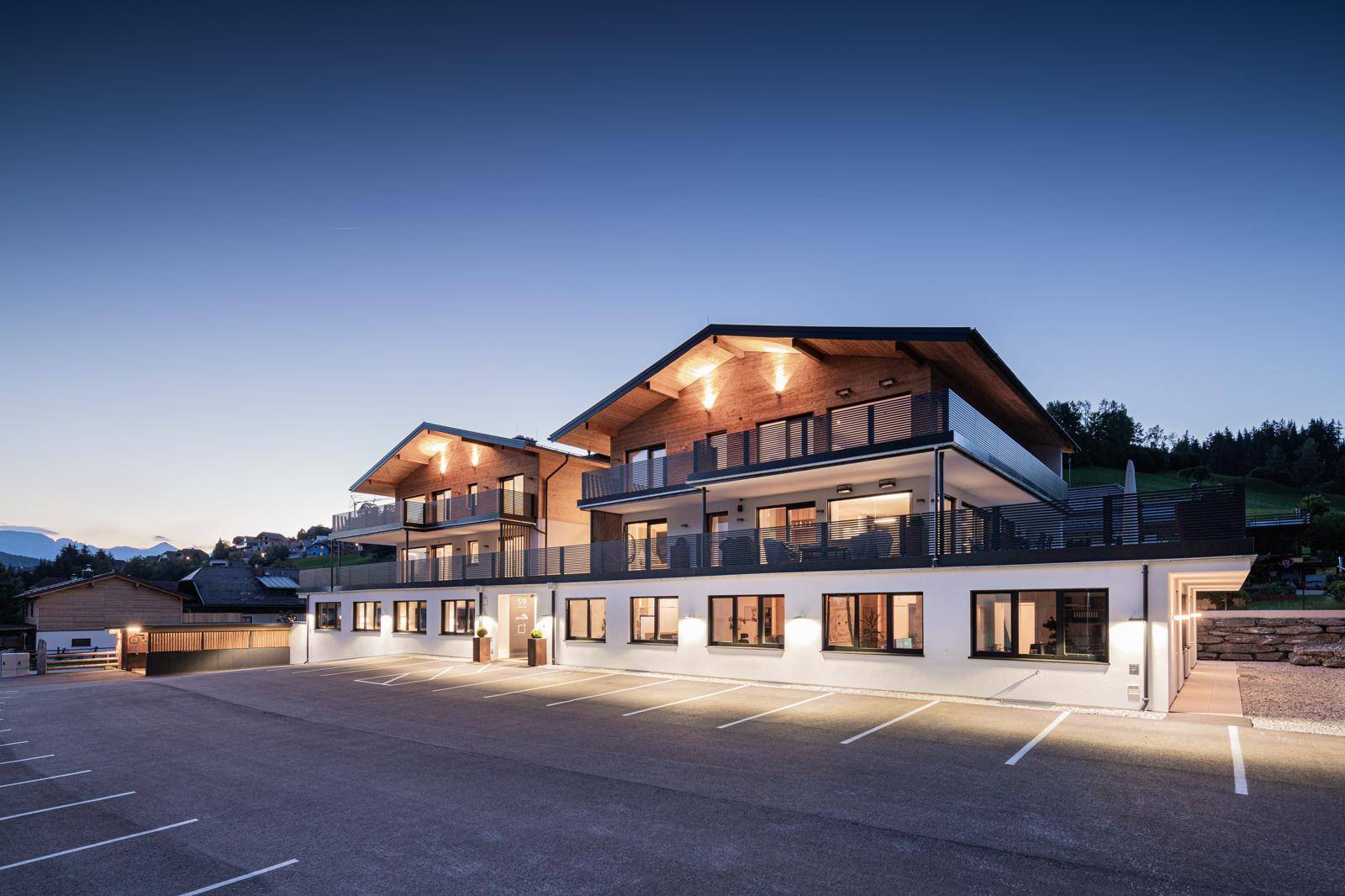 Modernes Wohngebäude bei Sonnenuntergang mit beleuchteter Fassade und Parkplätzen.