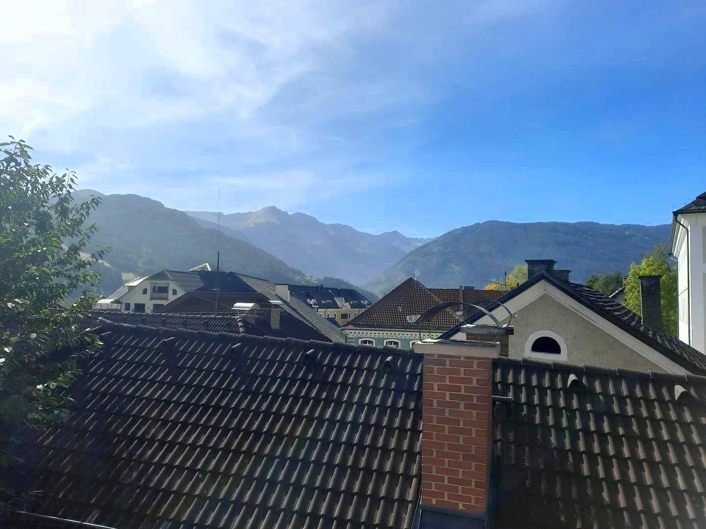 Weitläufiger Blick über die Dächer der Stadt auf die umliegende grüne Berglandschaft unter blauem Himmel.