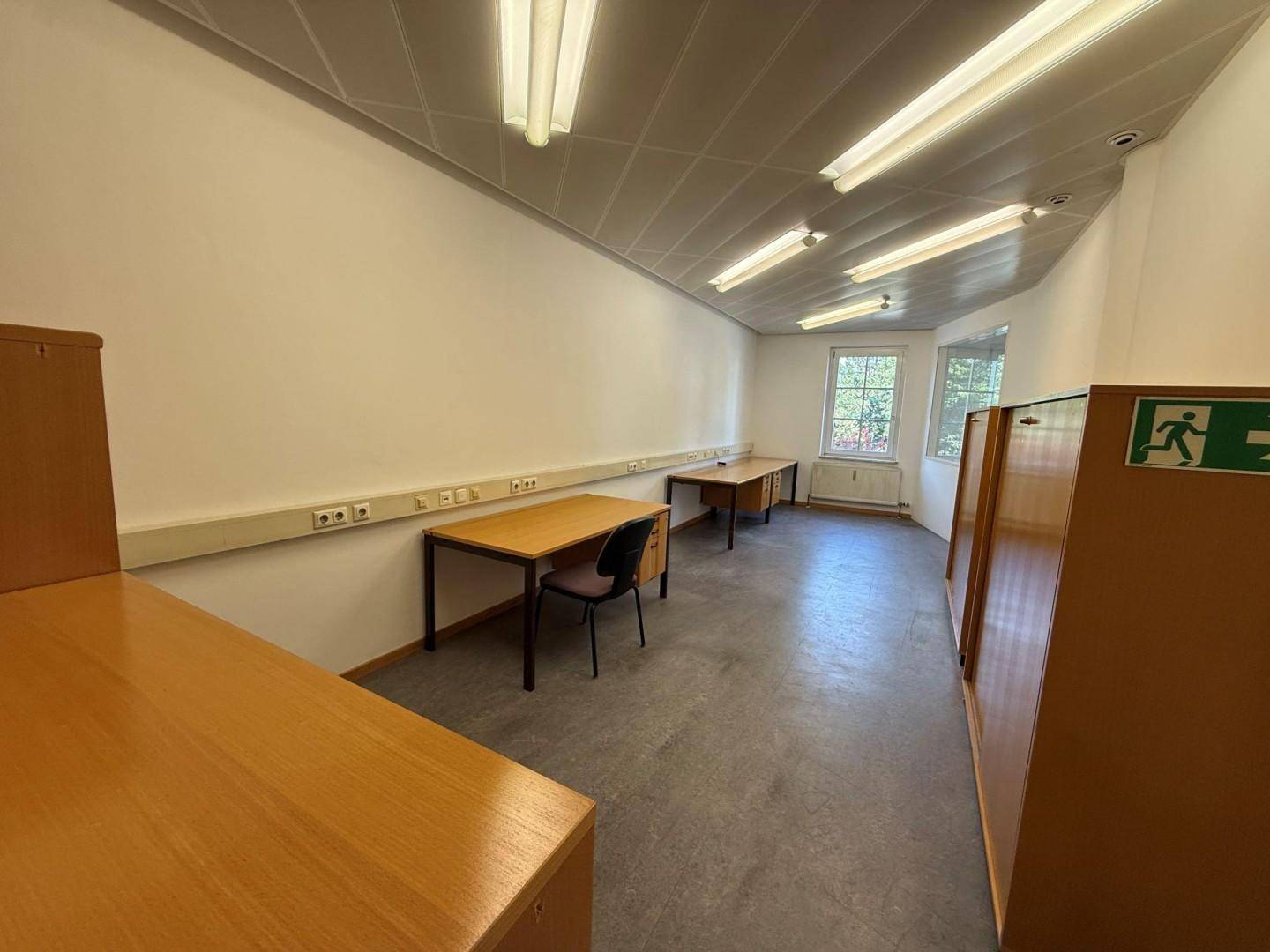 Langes Büro mit mehreren Schreibtischen entlang der Wand und Fenstern mit Tageslicht.