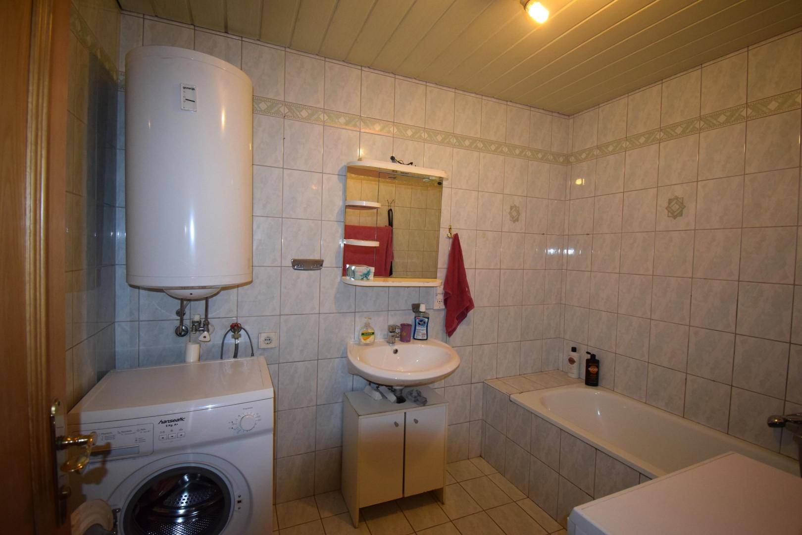 Badezimmer mit weißen Fliesen, Badewanne, Waschbecken und einem Warmwasserboiler.
