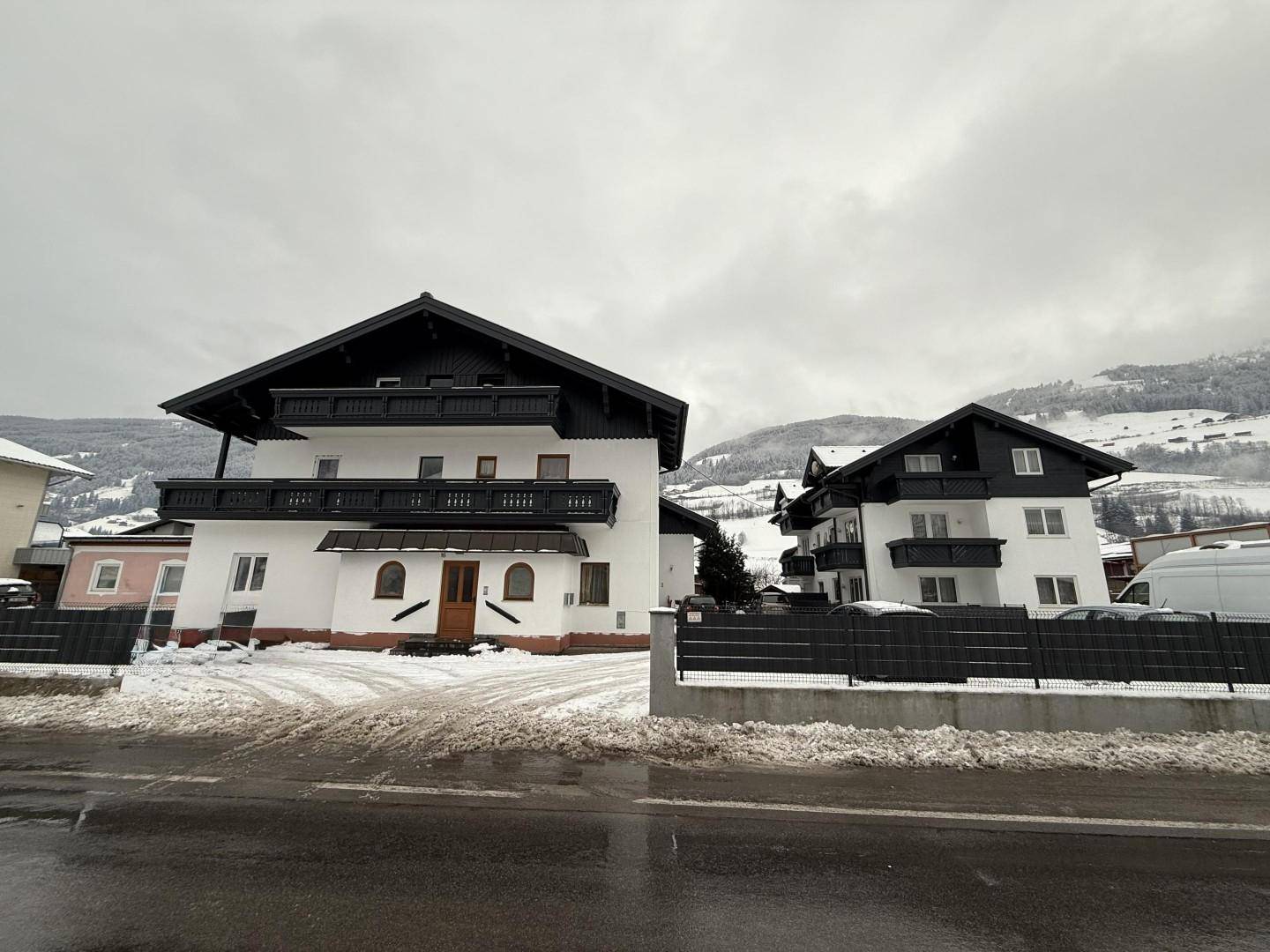 Zwei traditionelle Gebäude mit dunklen Dächern und Balkonen in verschneiter Berglandschaft.