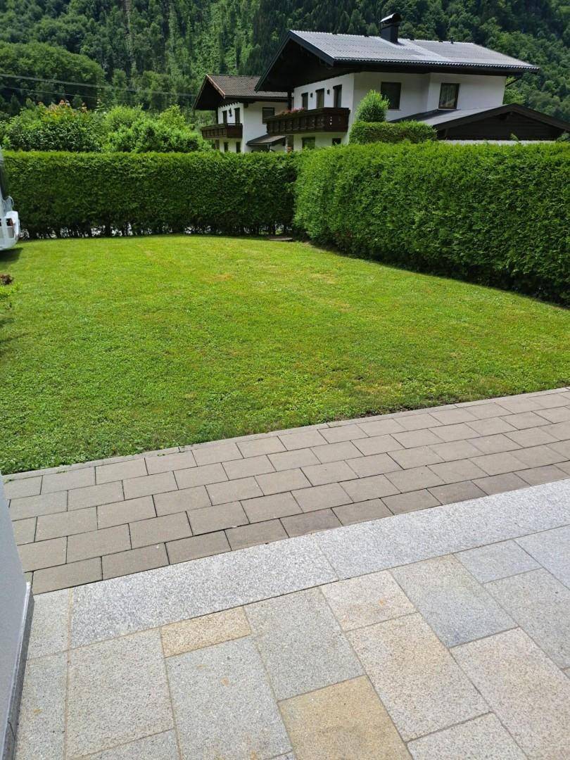 Gepflegte Rasenfläche mit Hecke und gepflastertem Weg vor einer Bergkulisse.