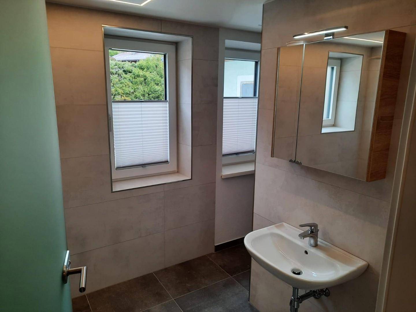 Helles Badezimmer mit modernem Waschbecken, Spiegelschrank und zwei Fenstern.