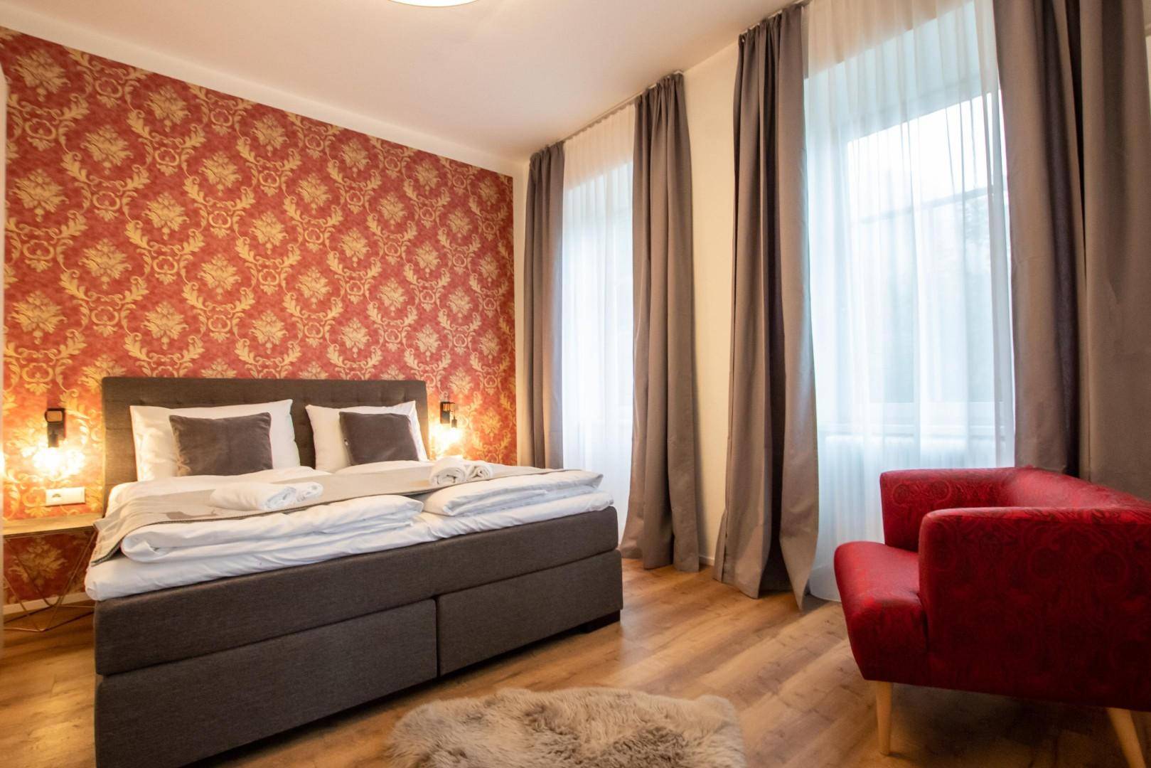 Schlafzimmer mit Doppelbett, rotem Sessel und auffälliger Tapete, hell beleuchtet.