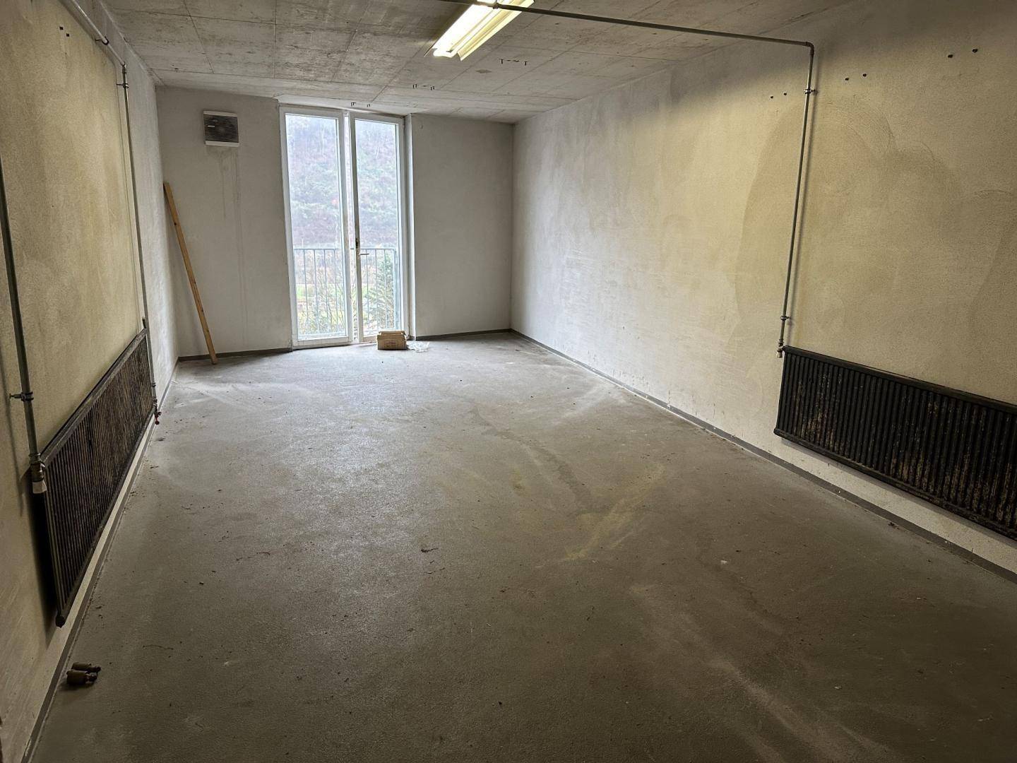 Großer Raum mit Betonboden, Heizkörpern und einer Fenstertür, die nach draußen führt.