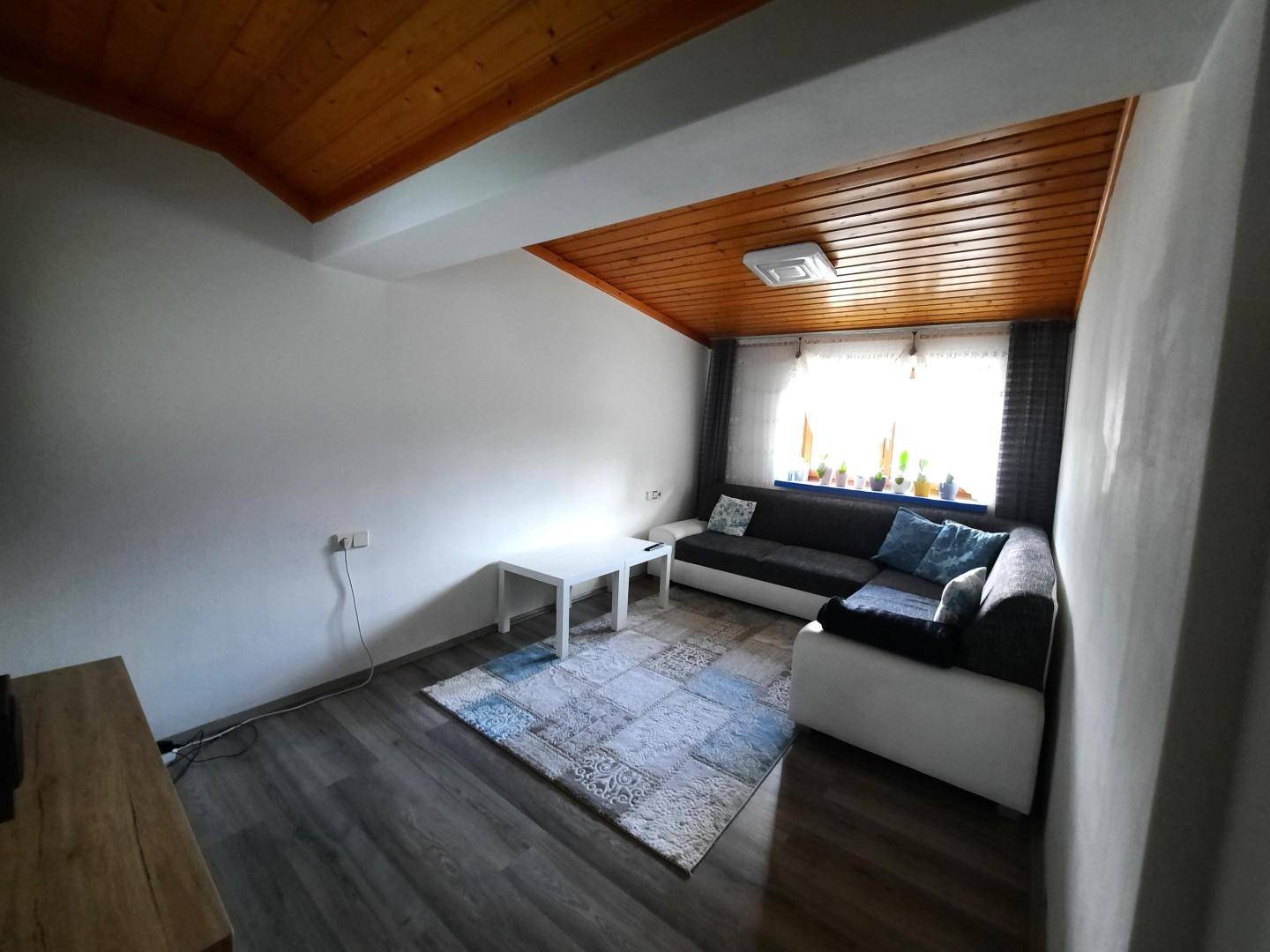 Gemütliches Wohnzimmer mit Holzbalkendecke, bequemer Couch und einem Fenster, das Tageslicht hereinlässt.