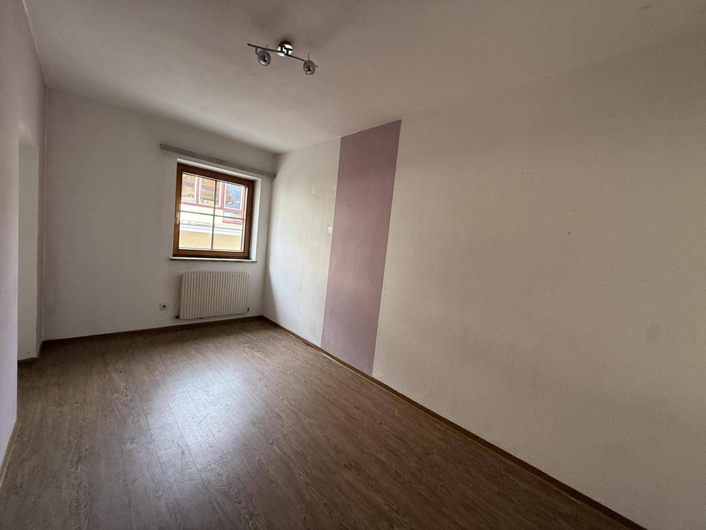 Leeres Zimmer mit Holzboden, einem Fenster und einer lila Akzentwand.