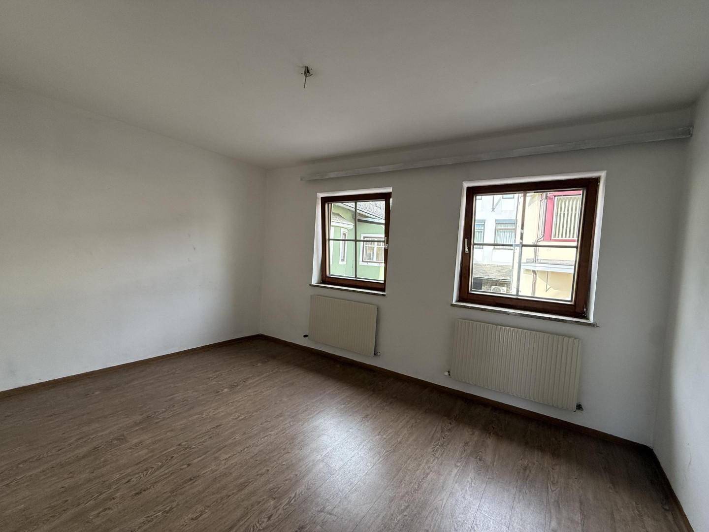 Helles Zimmer mit zwei Fenstern, Holzboden und weißen Wänden.