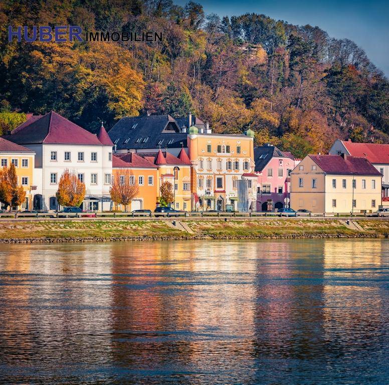 Malerische Flusslandschaft mit bunten Gebäuden am Ufer und herbstlicher Vegetation im Hintergrund.