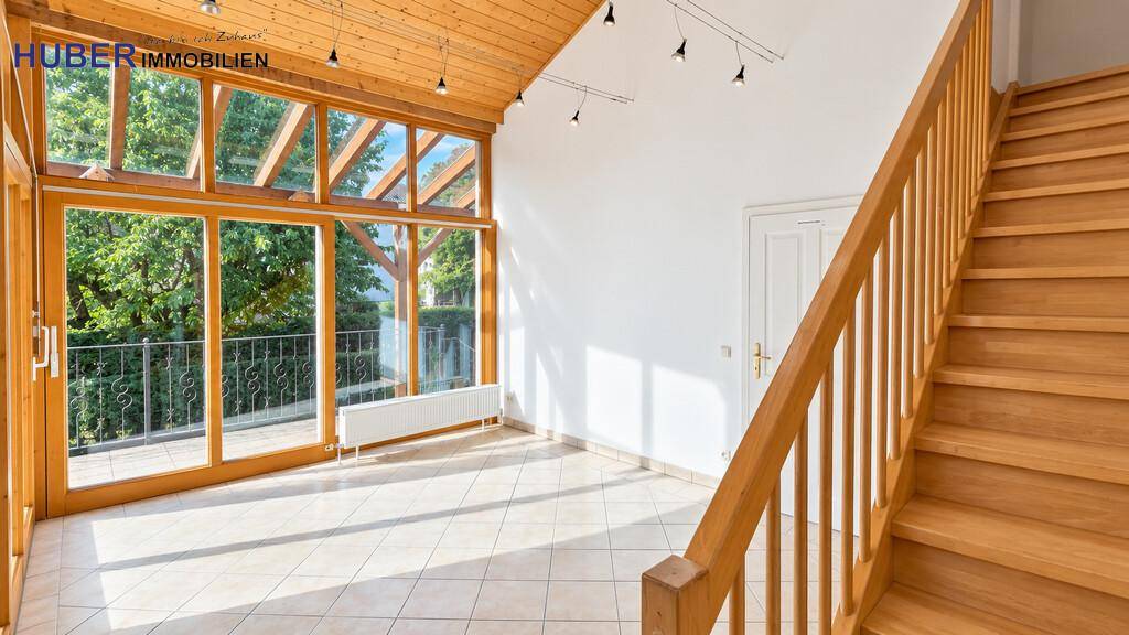 Sonniger Wintergarten mit großen Glasfronten, Fliesenboden und Holztreppe im Hintergrund.