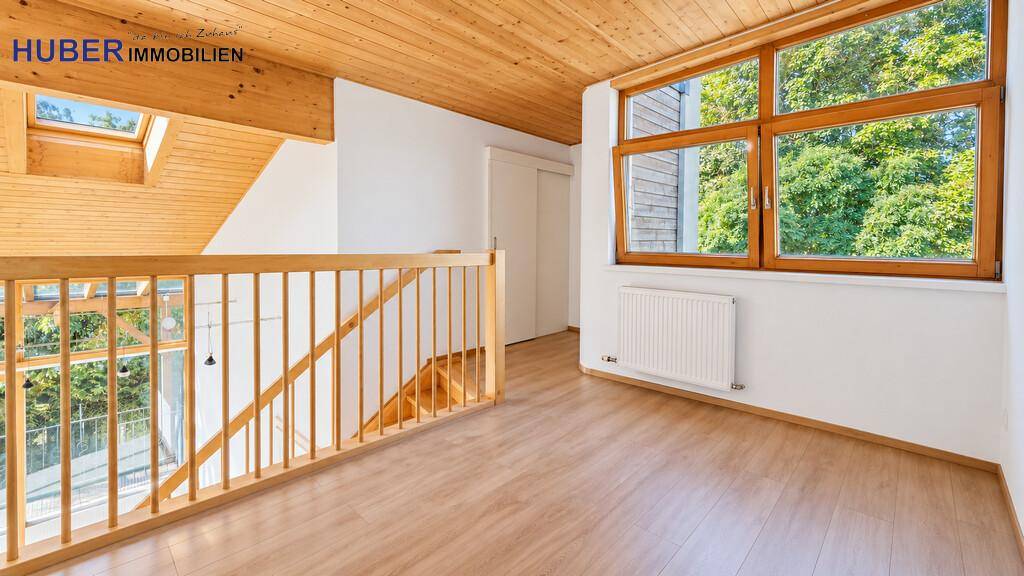 Heller Flur im Obergeschoss mit Holzdecke, Geländer und Blick ins Grüne durch große Fenster.