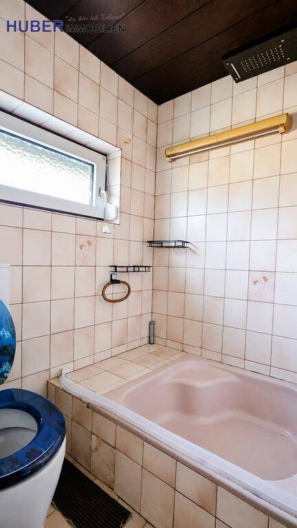 Badezimmer mit Badewanne, Duschkopf und Fenster, ausgestattet mit beigefarbenen Fliesen.