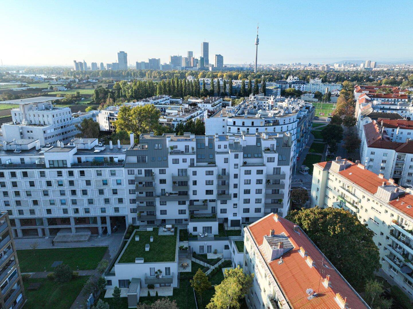 Weitläufige Luftaufnahme des Wohnkomplexes mit Stadtpanorama und grünen Dächern im Vordergrund.
