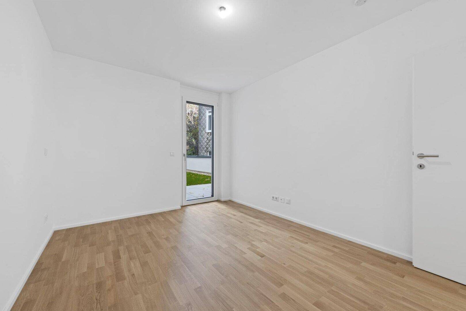 Leeres Schlafzimmer mit hellem Parkettboden und einer Tür, die zu einem Außenbereich führt.