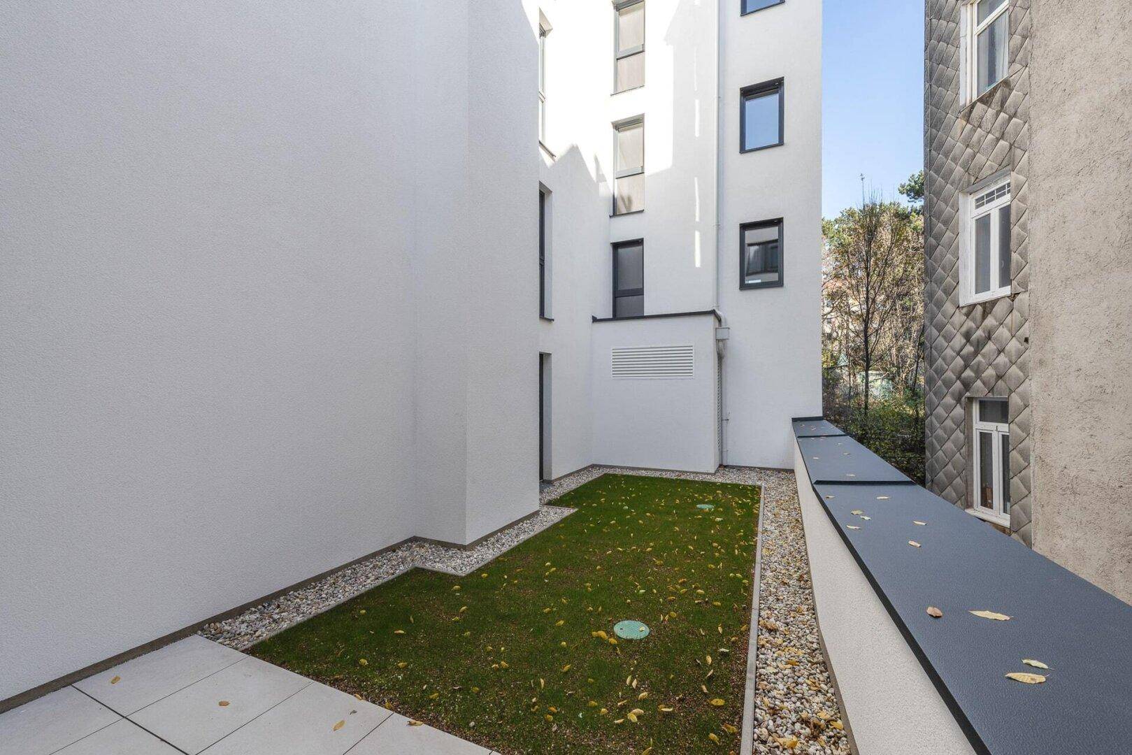 Kleiner, gepflegter Gartenbereich mit Rasenfläche und Kiesstreifen zwischen modernen Gebäudefassaden.