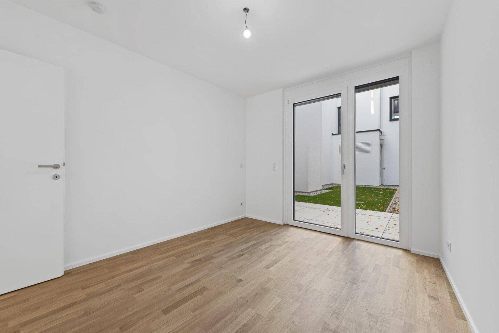 Helles Zimmer mit Parkettboden und großen Glastüren, die den Zugang zu einem kleinen Garten ermöglichen.