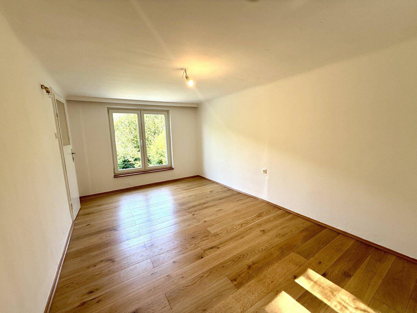Leeres Zimmer mit hellem Holzfußboden und einem großen Fenster mit Blick ins Grüne.