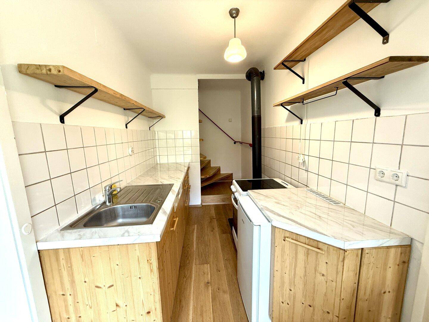 Funktionale Küche mit Holzschränken, weißen Fliesen und offenen Regalen, mit Treppenzugang.