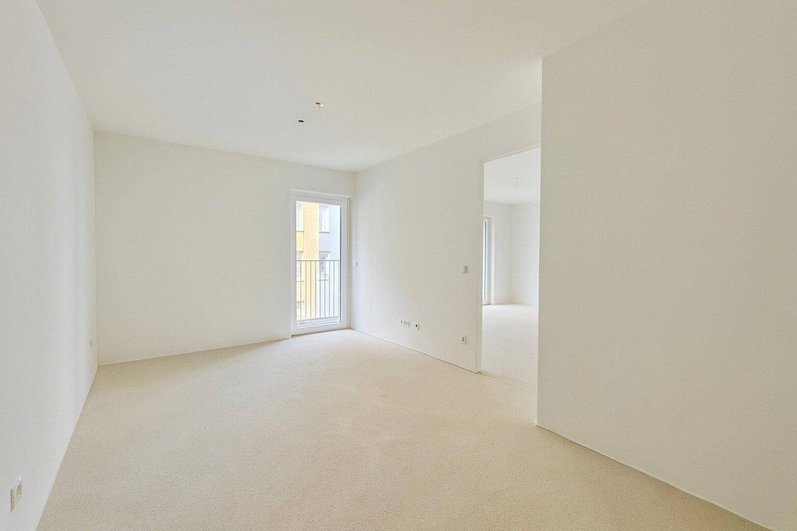 Heller, unmöblierter Wohnraum mit Teppichboden und Zugang zu einem kleinen Balkon.