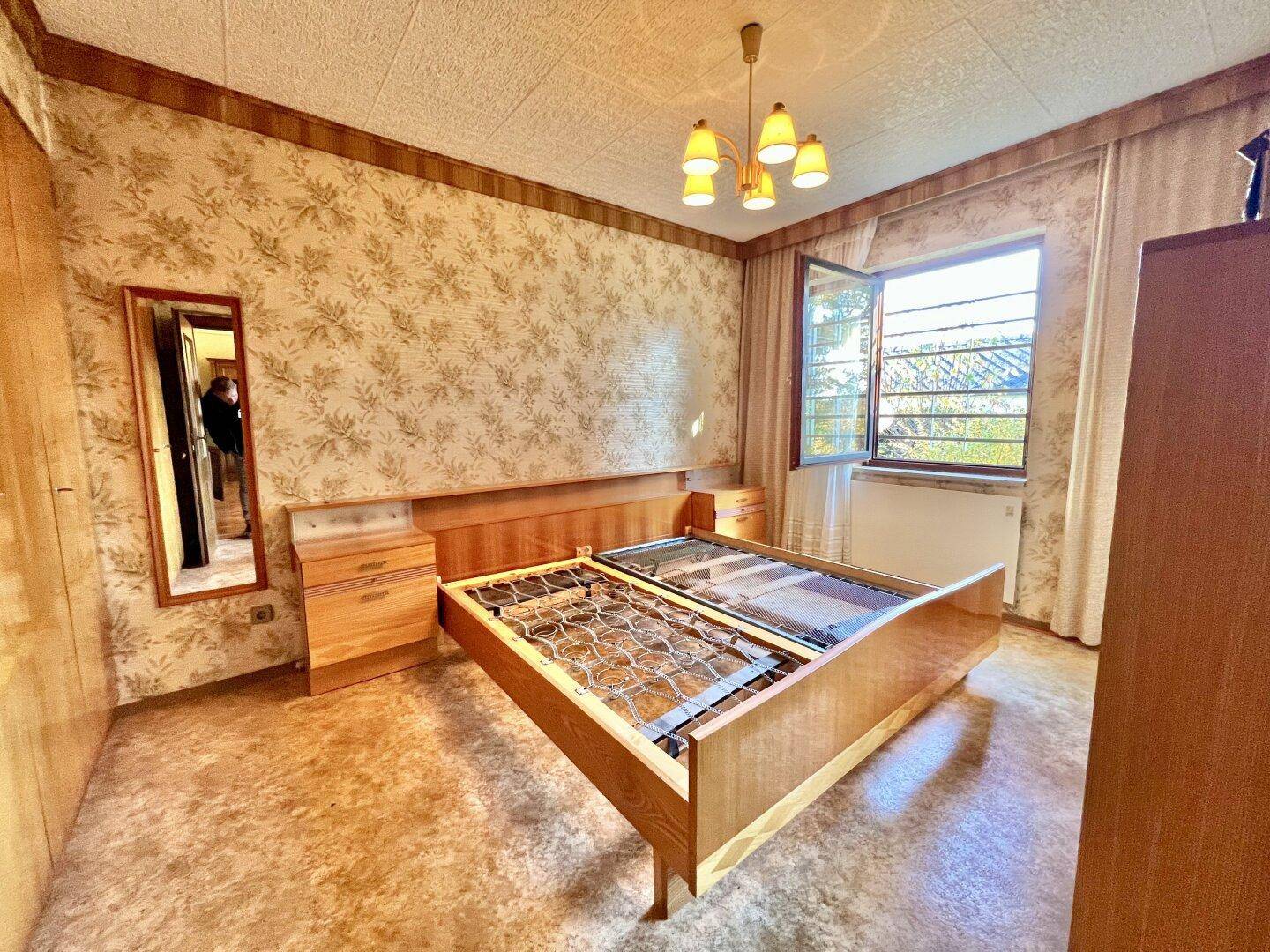 Schlafzimmer mit gemusterter Tapete, Holzmöbeln und einem Fenster für frische Luft und Tageslicht.