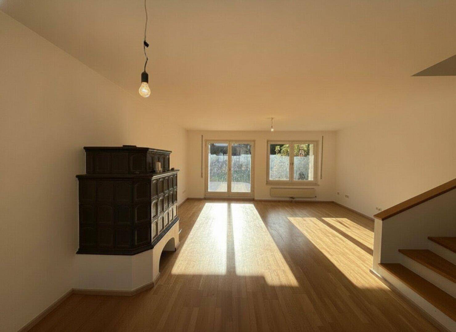 Lichtdurchflutetes Wohnzimmer mit Holzboden und traditionellem Kachelofen, unmöbliert.