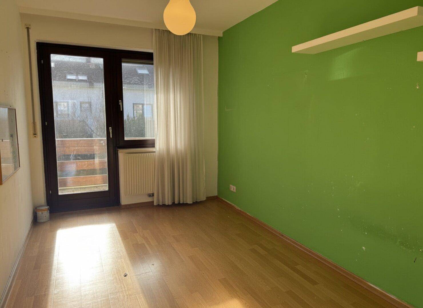 Leeres Kinderzimmer mit grün gestrichener Wand, Holzboden und Balkonzugang.