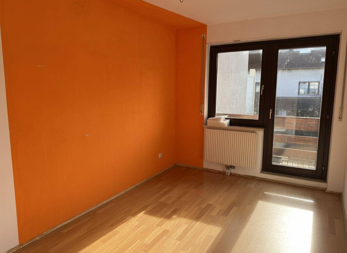 Leerer Raum mit orangefarbener Akzentwand, Holzboden und großem Fenster.
