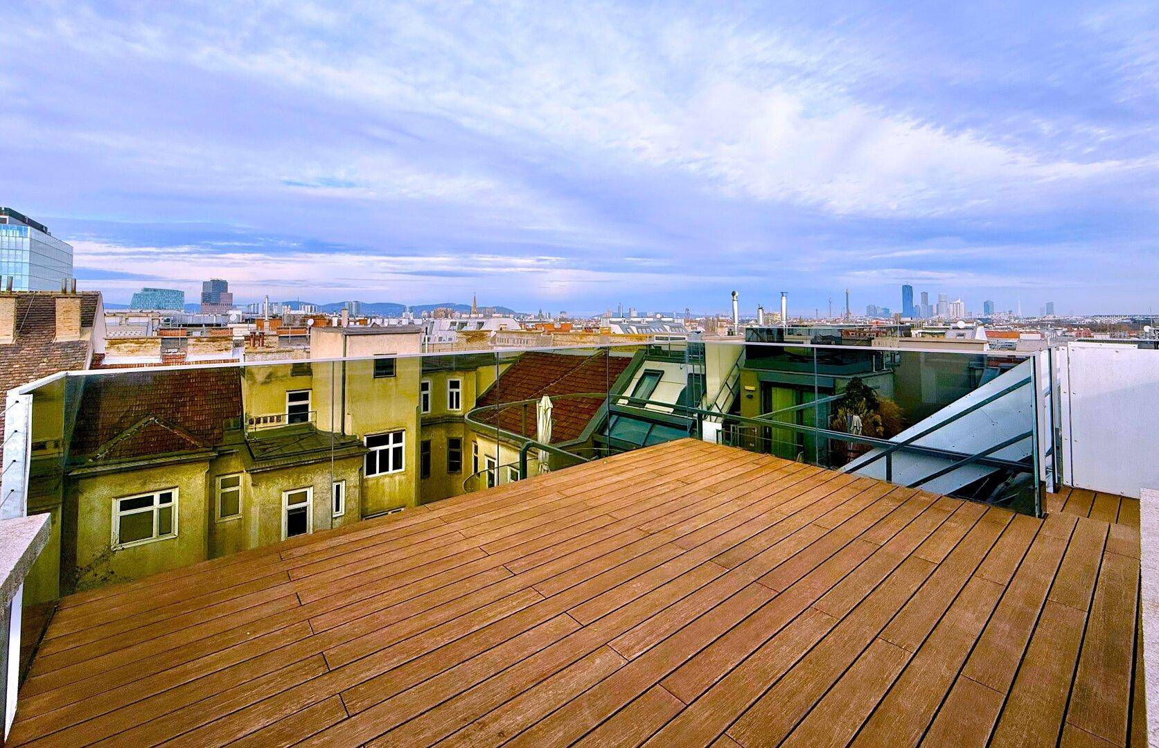 Geräumige Dachterrasse mit Holzplanken und Glasbrüstung, bietet eine weite Aussicht auf die städtische Umgebung.