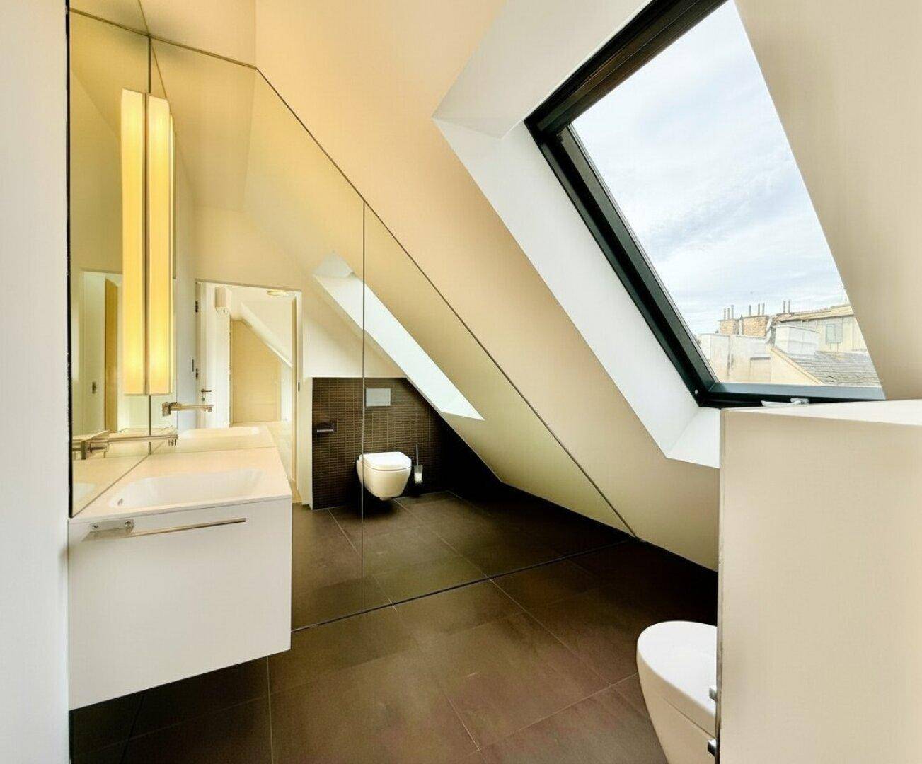 Modernes Badezimmer mit dunklen Fliesen, wandhängendem WC und Dachfenster, das viel Tageslicht hereinlässt.