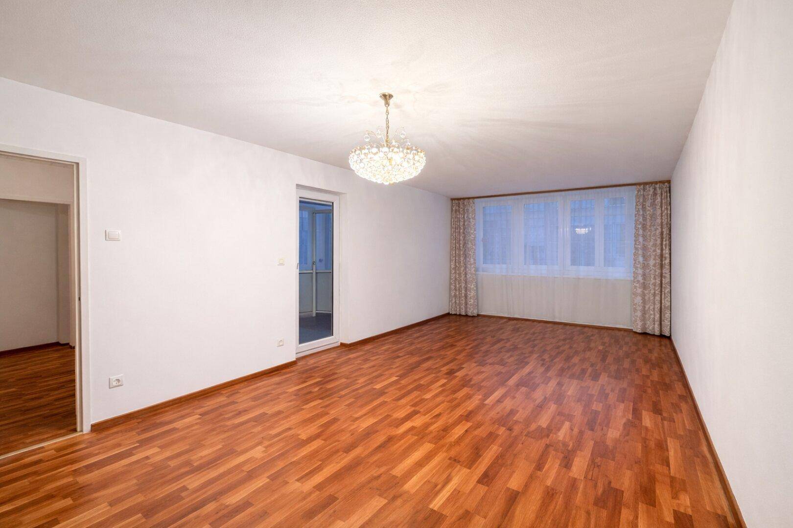 Geräumiger, leerer Wohn-Essbereich mit Holzboden, weißen Wänden und Balkonzugang.