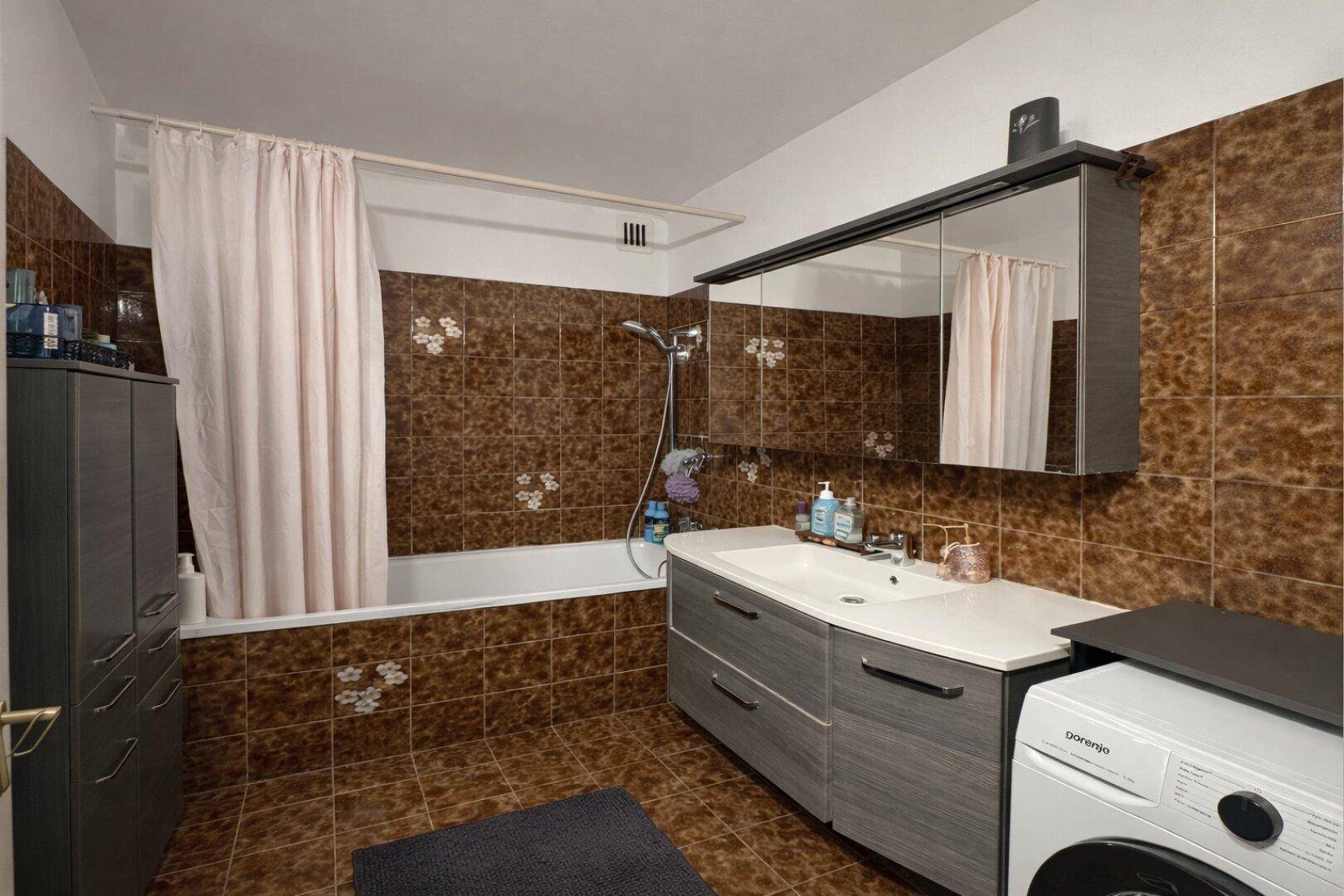 Badezimmer mit braunen Fliesen, Badewanne, Einzelwaschbecken und Waschmaschine.