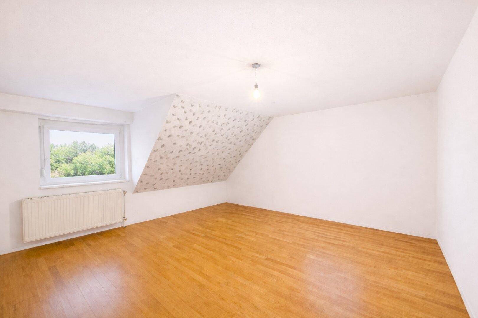 Geräumiges Schlafzimmer im Dachgeschoss mit Schrägen, Holzboden und Fenster, bereit zur Einrichtung.