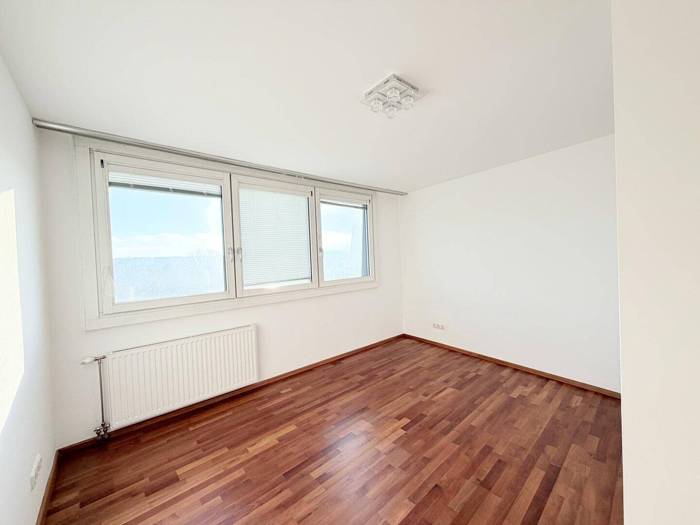 Leeres Zimmer mit Holzboden, Heizkörper und großen Fenstern mit Jalousien.