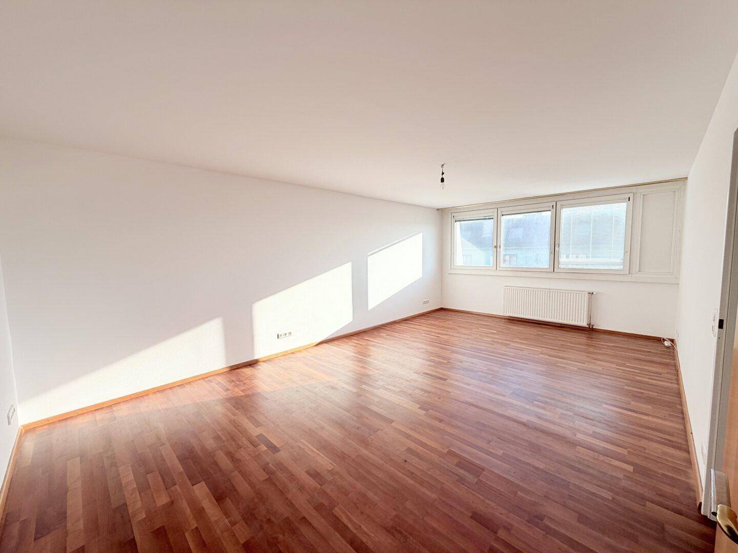 Heller, leerer Wohnbereich mit Holzboden und großen Fenstern, die viel Sonnenlicht hereinlassen.