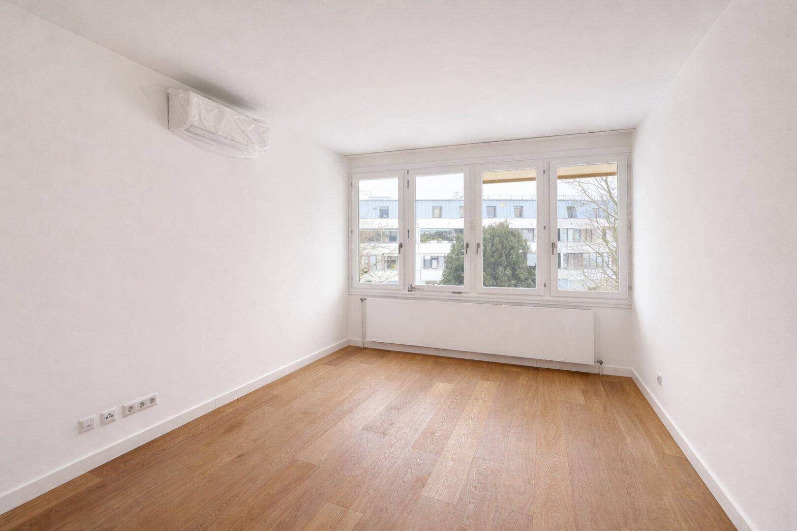 Leeres Zimmer mit Holzboden, großen Fenstern und Klimaanlage.