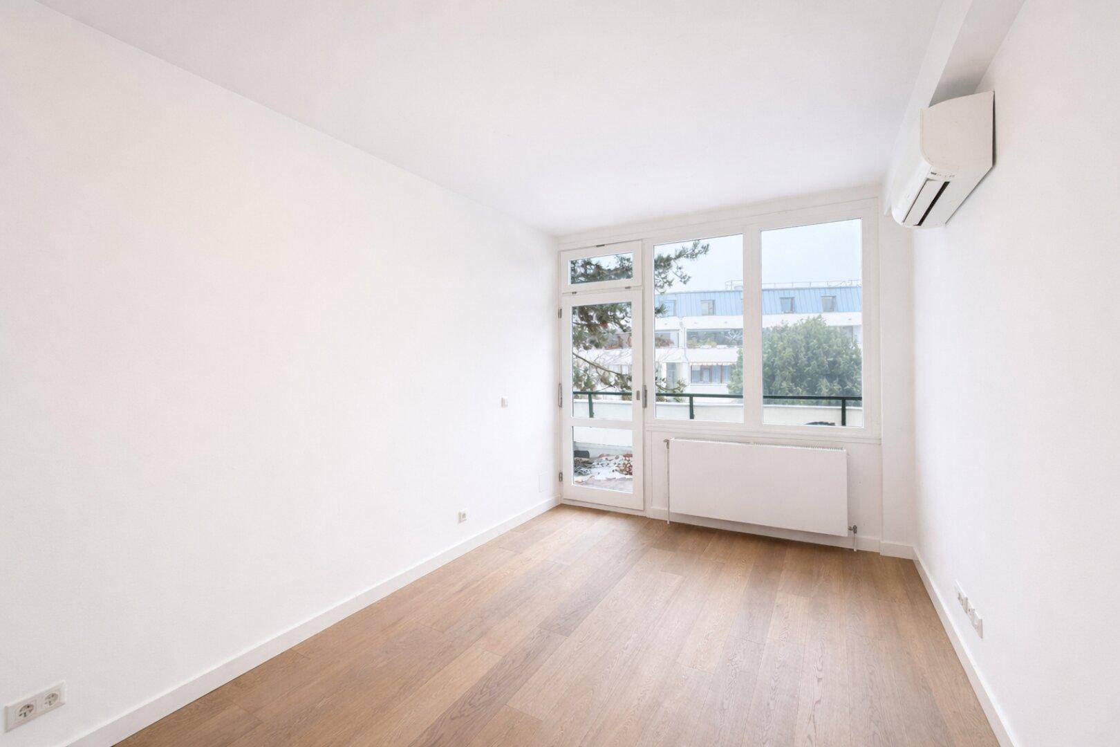 Leeres Zimmer mit Holzboden, großen Fenstern und Klimaanlage.