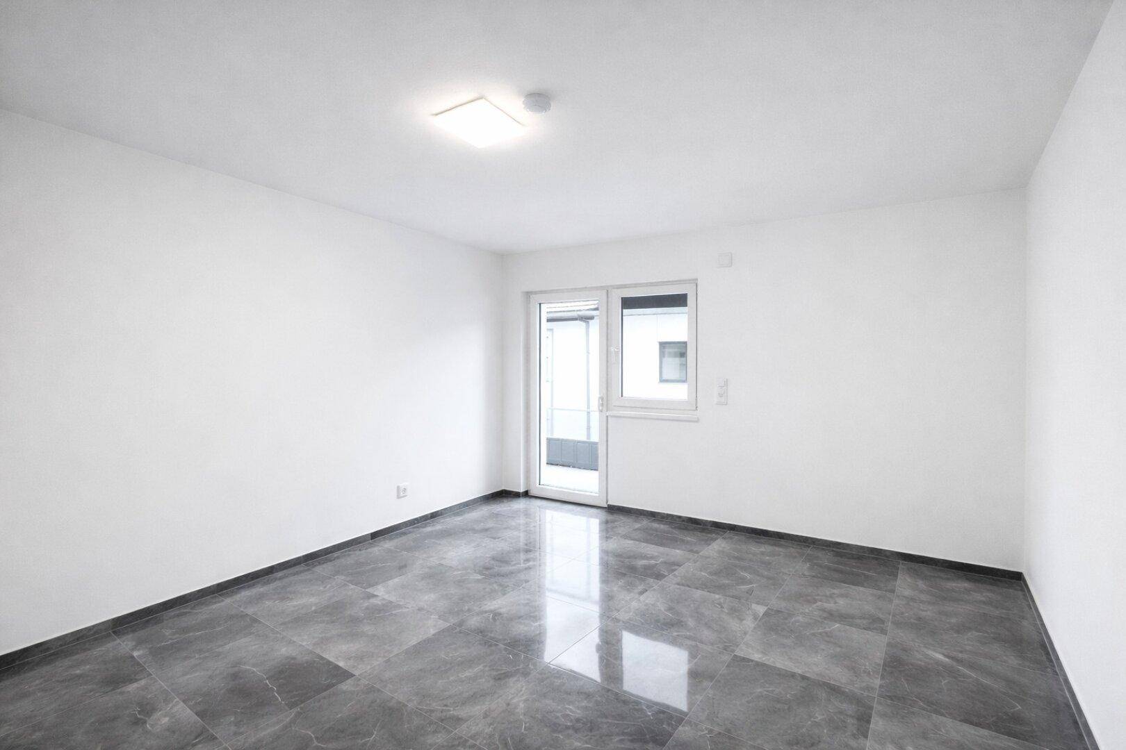 Leeres Zimmer mit Fliesenboden und Fensterfront, die zu einem Außenbereich führt.
