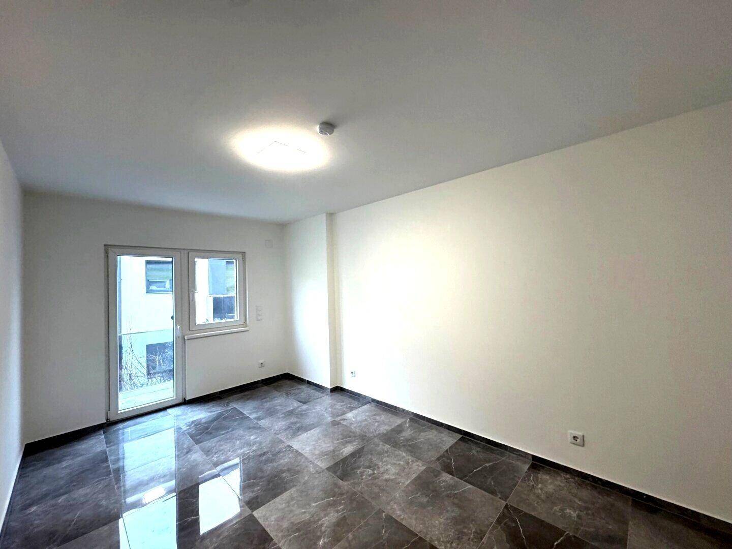 Leeres Zimmer mit Fliesenboden und Fensterfront, die zu einem Außenbereich führt.